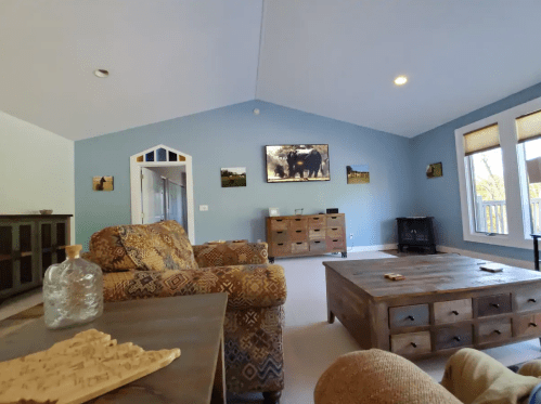 Cozy living room with blue walls, patterned furniture, a wooden coffee table, and large windows letting in natural light.