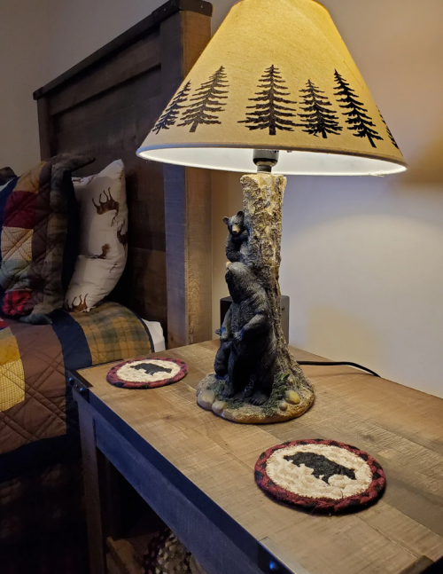 A rustic lamp with a bear design sits on a wooden nightstand, next to two round coasters, in a cozy bedroom setting.