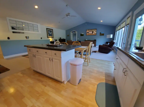 Bright, open kitchen and living area with wooden floors, white cabinets, and large windows. Cozy seating in the background.