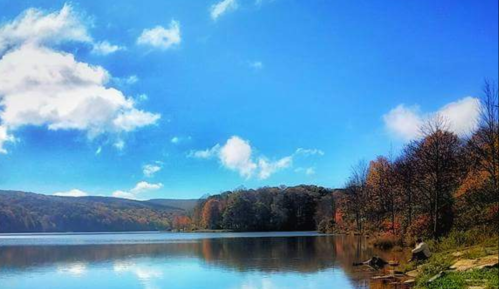 A serene lake surrounded by autumn trees under a bright blue sky with fluffy clouds. Reflections shimmer on the water.