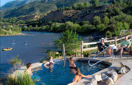 People relax in a hot spring by a river, with kayakers and paddleboarders in the background and mountains in the distance.