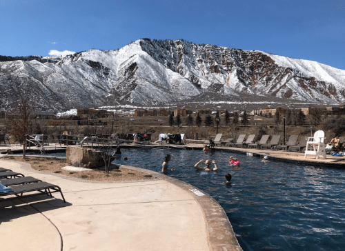 A scenic outdoor pool surrounded by mountains, with people swimming and lounging on deck chairs under a clear blue sky.