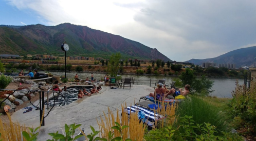 A scenic riverside area with people relaxing on lounge chairs, surrounded by mountains and greenery.