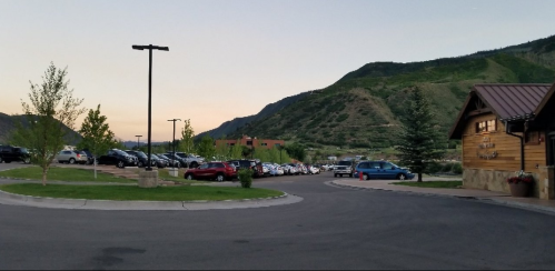 A parking lot with numerous cars, surrounded by green hills and a wooden building at sunset.