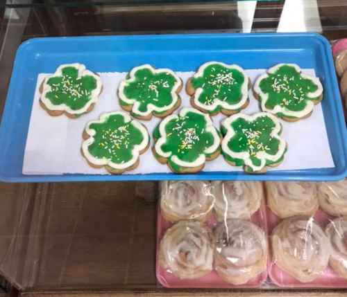 A tray of green shamrock-shaped cookies with white icing and sprinkles, displayed in a bakery.