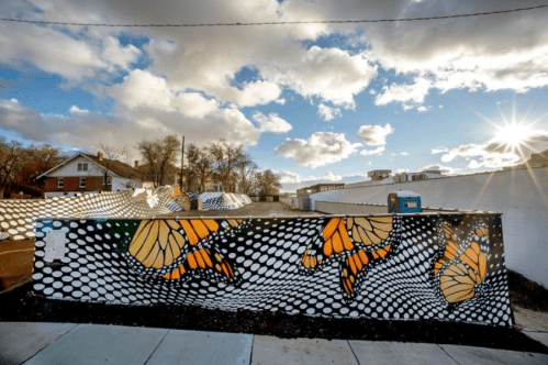 Colorful mural of butterflies on a wall, with a bright sky and clouds in the background. Sunlight shines in the corner.