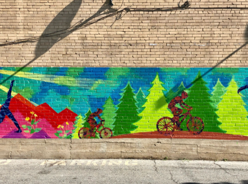 Colorful mural depicting cyclists and trees against a vibrant landscape of mountains and flowers on a brick wall.
