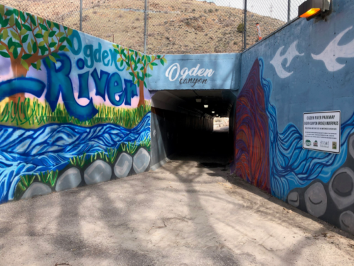 Colorful mural at the entrance of a tunnel, featuring "Ogden River" and scenic designs, leading into a dark passageway.