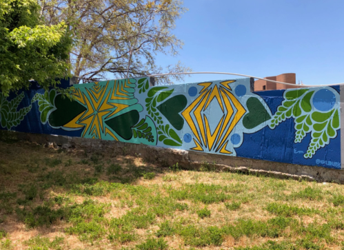 Colorful mural featuring geometric shapes and leafy patterns on a blue wall, surrounded by green grass and trees.