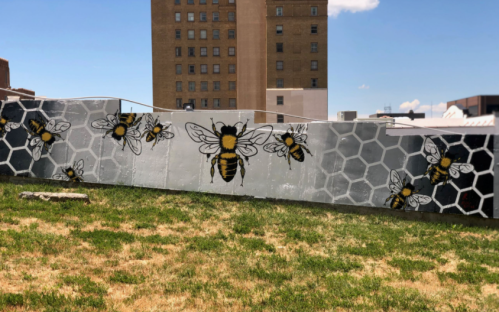 A mural featuring bees on a honeycomb pattern, set against a grassy area and urban buildings in the background.
