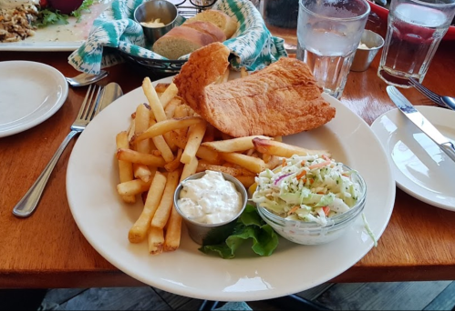 A plate with crispy fried fish, French fries, coleslaw in a cup, and a side of dipping sauce on a wooden table.