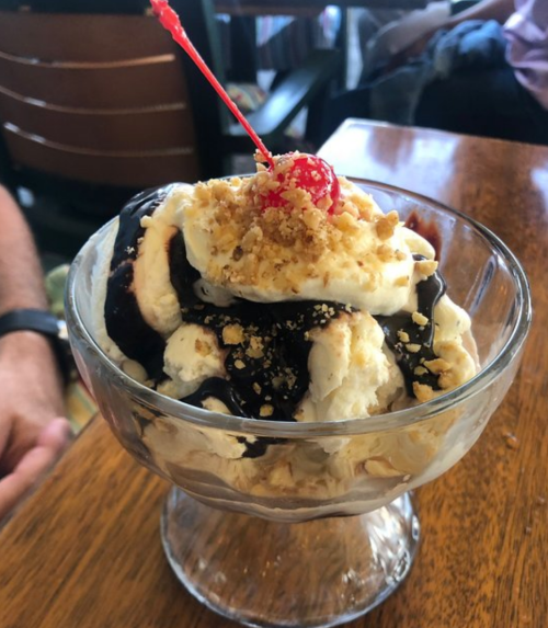 A glass dessert bowl filled with ice cream, chocolate sauce, crushed nuts, and topped with a cherry.