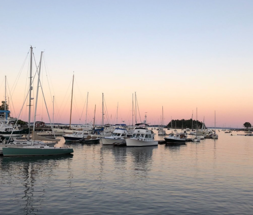 A serene harbor at sunset, with various boats anchored and a soft pastel sky reflecting on the calm water.