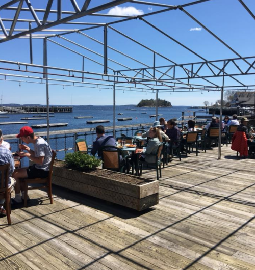 Outdoor dining area with people enjoying meals by the water, under a blue sky with boats in the background.