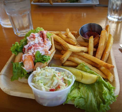 A plate with a lobster roll on lettuce, fries, coleslaw in a cup, and a pickle slice, served on a wooden tray.
