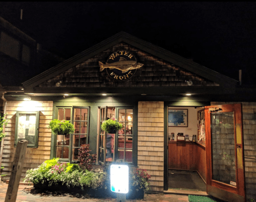 A cozy restaurant entrance at night, featuring wooden shingles, green trim, and illuminated signage.
