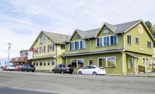 A green two-story building with multiple windows and parked cars in front, set against a cloudy sky.