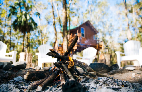 A campfire with burning sticks in the foreground, surrounded by rocks, with a treehouse and chairs in the background.
