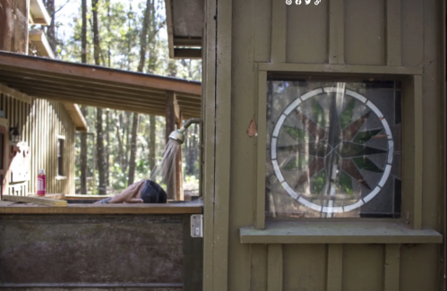 A rustic cabin with a stained glass window, featuring a shower outside and a person relaxing in the background.