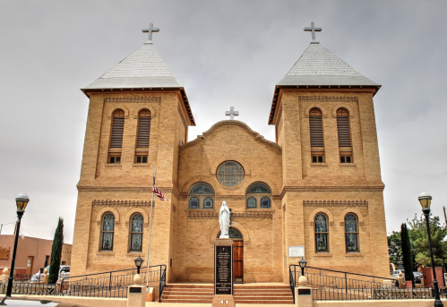 Historic brick church with two towers, crosses on top, and a statue in front, set against a cloudy sky.
