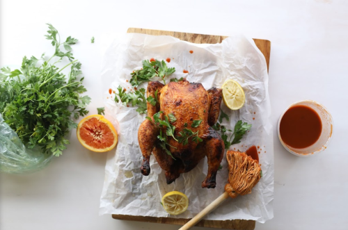 A roasted chicken garnished with herbs and lemon, accompanied by a bowl of sauce and fresh greens on a wooden board.