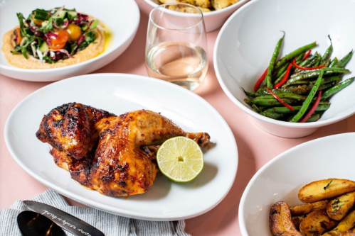 A plate with grilled chicken, lime, and sides of green beans, salad, and roasted potatoes, with a glass of white wine.