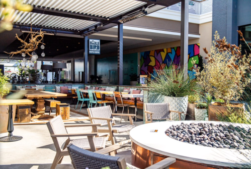 Outdoor dining area with modern furniture, colorful mural, and a fire pit surrounded by plants. Bright and inviting atmosphere.