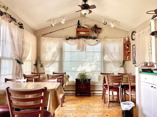 Cozy dining area with wooden tables, chairs, and large windows adorned with curtains and decorative items.