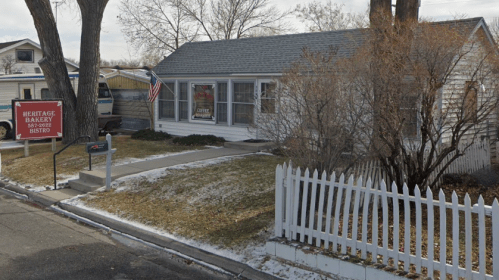 A small white house with a gray roof, featuring a bakery sign and a white picket fence, surrounded by sparse landscaping.