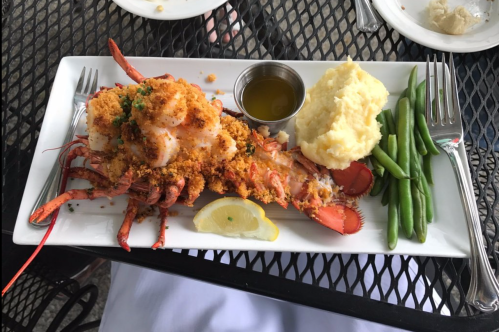 A plate featuring a baked lobster with breadcrumbs, mashed potatoes, green beans, and a lemon wedge.