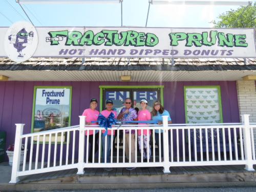 A group of five people stands in front of a colorful donut shop named "Fractured Prune," smiling and holding a ribbon.