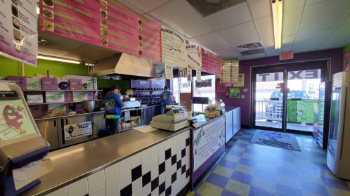 Interior of a colorful eatery with a counter, menu boards, and two customers at the register. Exit door visible.
