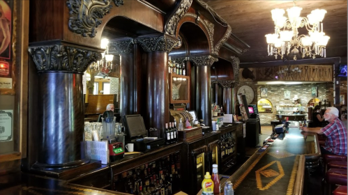 A vintage bar with ornate wooden columns, mirrors, and a well-stocked counter, featuring chandeliers and patrons.
