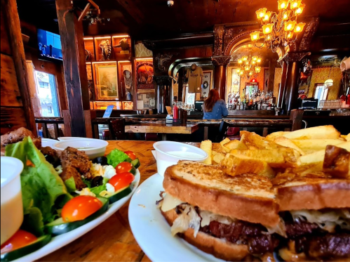 A cozy restaurant scene featuring a plate of sandwiches, fries, and a salad, with a bar and patrons in the background.