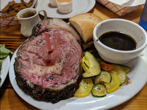 A plate featuring a large slice of prime rib with a red marker, served with zucchini, a roll, and a bowl of gravy.