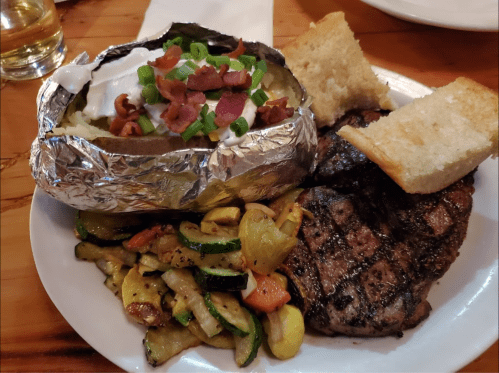 A plate featuring a grilled steak, baked potato with sour cream and bacon, and sautéed vegetables, with bread on the side.