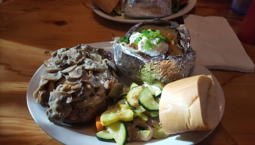 A plate featuring a mushroom-covered burger, a baked potato with toppings, sautéed zucchini, and a roll.