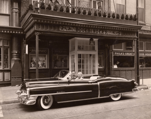 A vintage black convertible parked outside a historic building with "Bookbinders" signage. Two people are inside the car.
