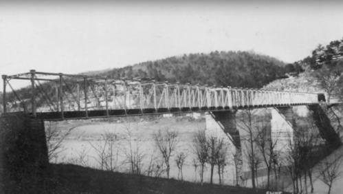 Black and white photo of a historic bridge spanning a river, surrounded by hills and bare trees.