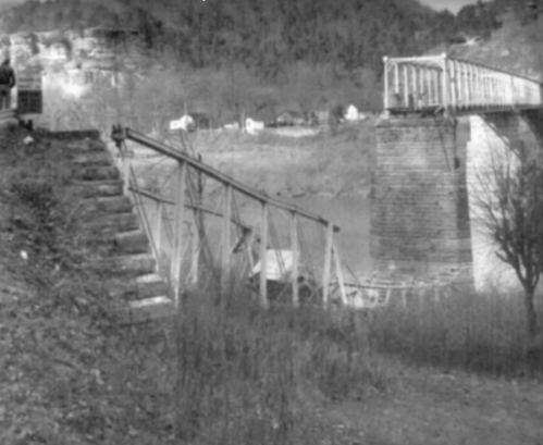 Black and white image of a dilapidated bridge with wooden supports and stone steps leading to the riverbank.