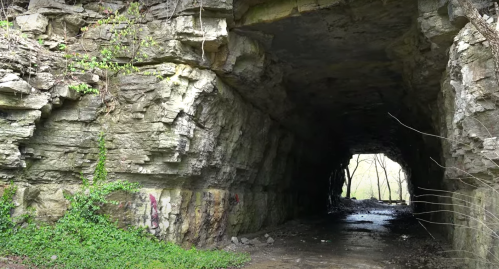 A rocky tunnel with greenery and graffiti, leading into a bright, open area at the far end.