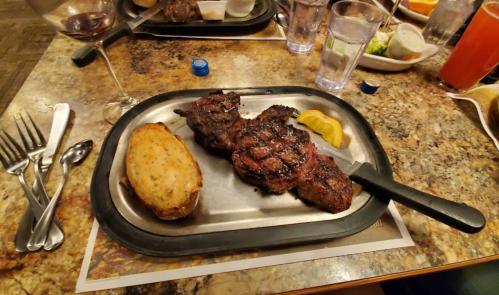 A sizzling steak dinner on a metal plate, accompanied by a baked potato and lemon slice, with drinks in the background.