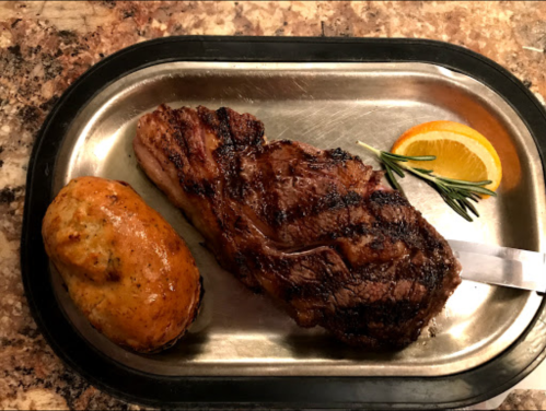 A grilled steak on a metal plate, accompanied by a baked potato, an orange slice, and a sprig of rosemary.