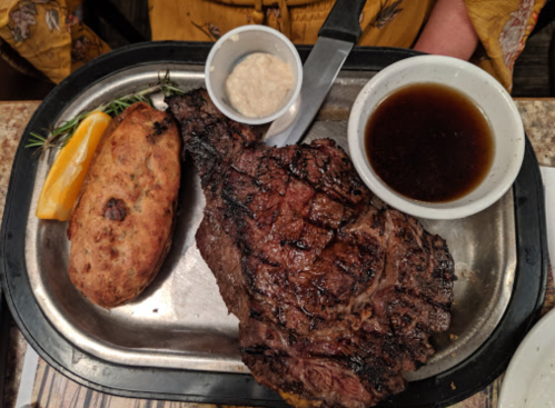 A grilled ribeye steak on a metal plate, accompanied by a baked potato, dipping sauces, and a lemon wedge.