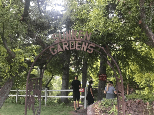 A metal archway reading "Sunken Gardens" leads into a lush, green garden area with visitors walking underneath it.