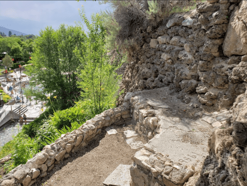 Stone steps lead down a rocky hillside, surrounded by greenery, with a glimpse of a pool area in the background.