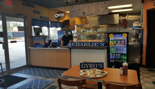 A casual eatery interior with a counter, menu board, and a table with gyros. A staff member is at the counter.