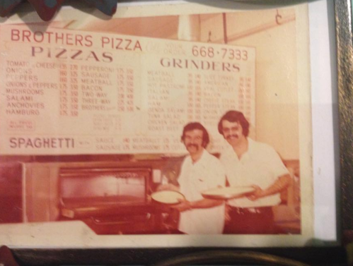 Two men stand in front of a pizza menu, holding pizza trays, smiling in a vintage pizzeria setting.