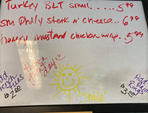Menu board with food items and prices, including a Turkey BLT, Philly steak, and a cheerful sun drawing.