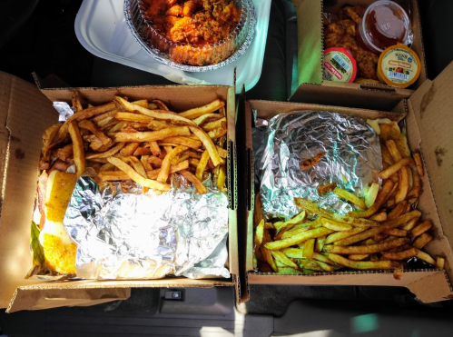 Two takeout boxes filled with golden fries, one with a foil-wrapped item, and a container of fried chicken in the background.